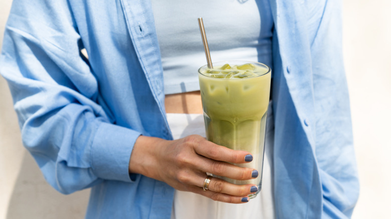 Woman holding an iced matcha latte in front of her light-colored outfit.