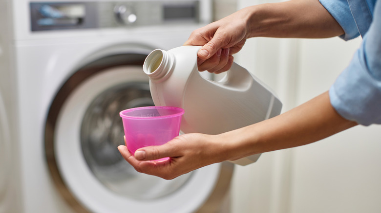 Someone pouring laundry detergent into a plastic cup with a washing machine in the background
