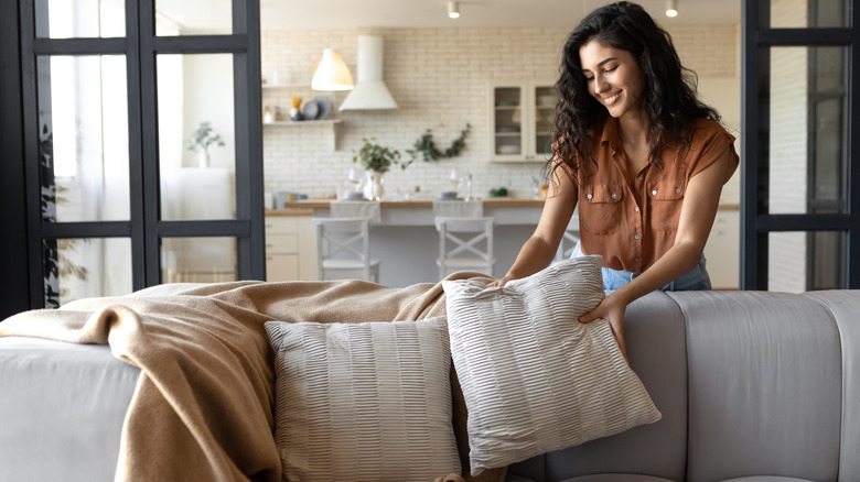 Young woman putting soft pillows and plaid on comfy sofa, making her home cozy and warm.