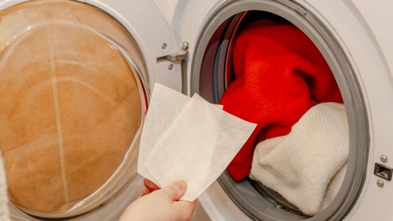 A hand holding color catcher sheets in front of a front-loading washing machine filled with red and white laundry to prevent dye transfer during the wash cycle.