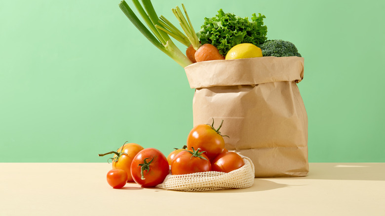 Tomatoes sitting on a counter on top of a net bag and next to a paper bag filled with vegetables