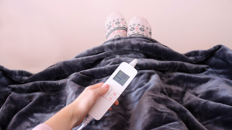Someone under dark colored electric blanket holding controler in hand with slippered feet showing out the bottom
