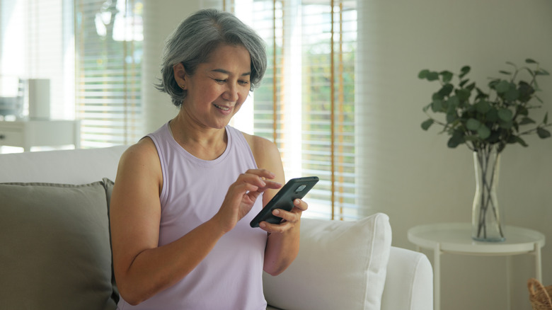 An Asian woman sits on a white sofa next to a vase full of eucalyptus while using a smartphone