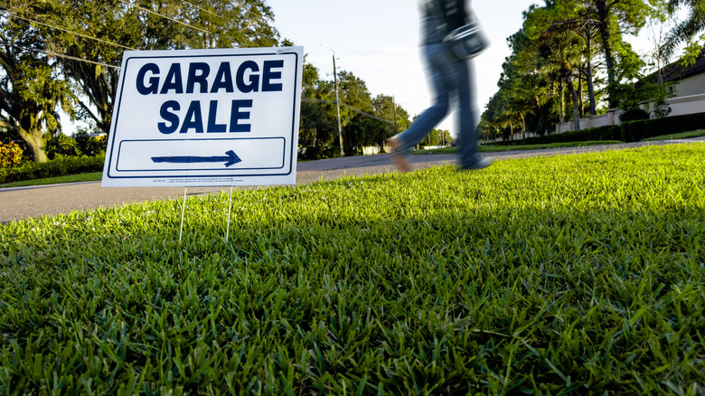 Garage sale sign in green yard with blurred person walking past
