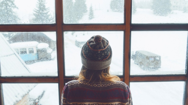 Person in sweater and hat standing in front of window looking out at snowy drive