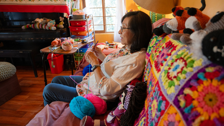 A woman crocheting on her couch with yarn and finished projects all around her