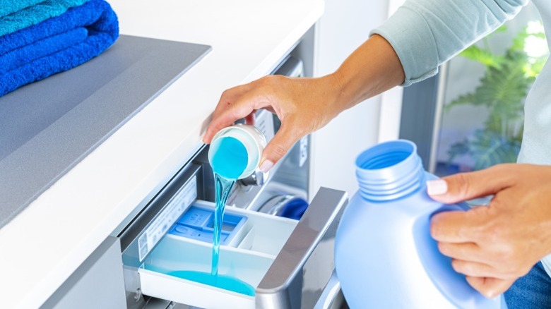 Person pouring blue liquid laundry detergent into washing machine drawer.