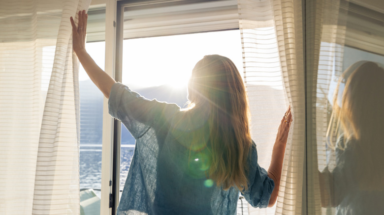 Woman opening white curtains in front of sliding glass door.