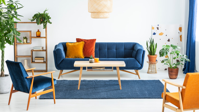 A living room with a blue sofa and chair, orange pillows and chair, with a bookcase and plants in the background.