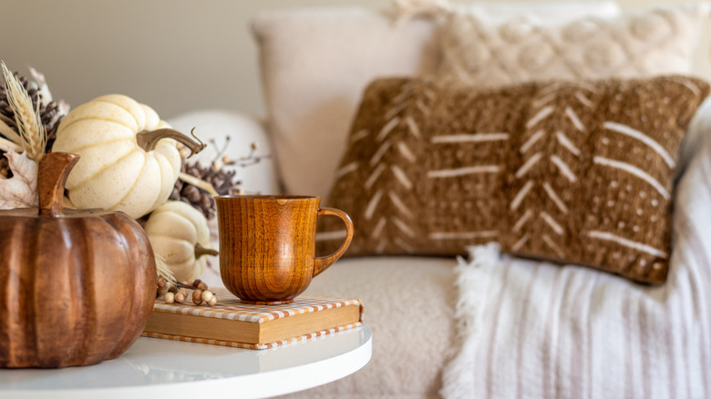 A living room space with white and brown pumpkins alongside dried plants with a wooden cup and a book being used as a coaster in the front