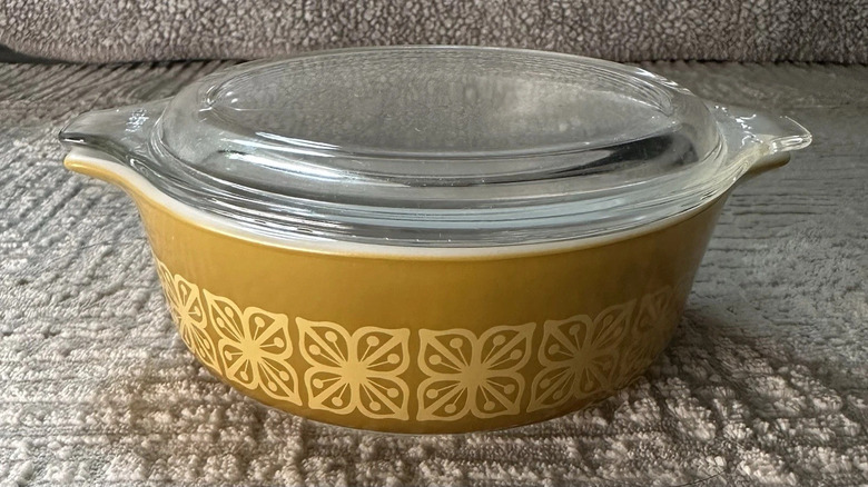 A mustard-colored vintage Pyrex glass-lidded casserole dish adorned with the Autumn Floral pattern sits on a fabric couch.