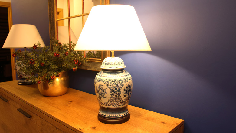 A blue and white flower patterned lamp sitting on a sideboard with dim lighting and a blue wall.