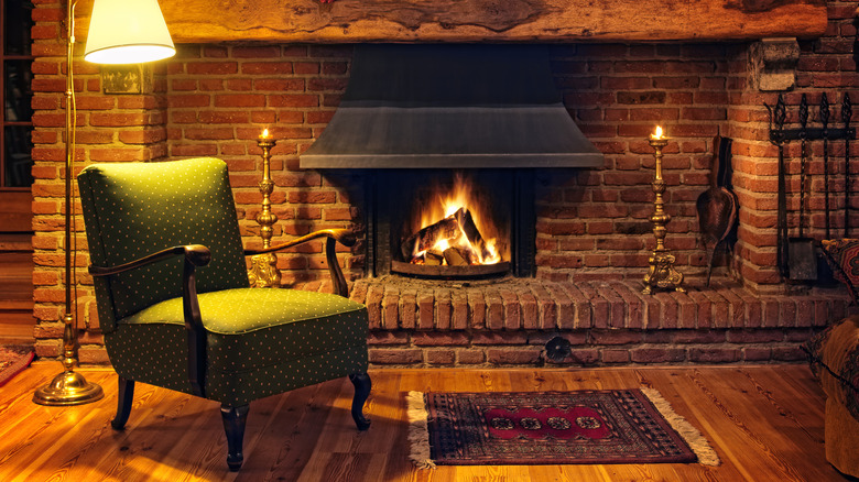 An older brick fireplace in a cozy living room with a green armchair, a small rug, and a tall floor lamp