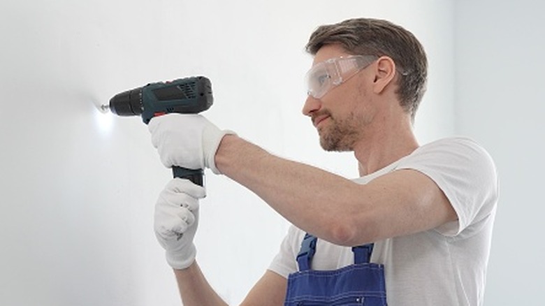 A man in goggles and blue overalls drills into a white sheet of drywall.