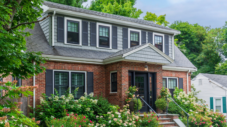 The outside of an older home with many windows and a black front door surrounded by trees and lush floral landscaping