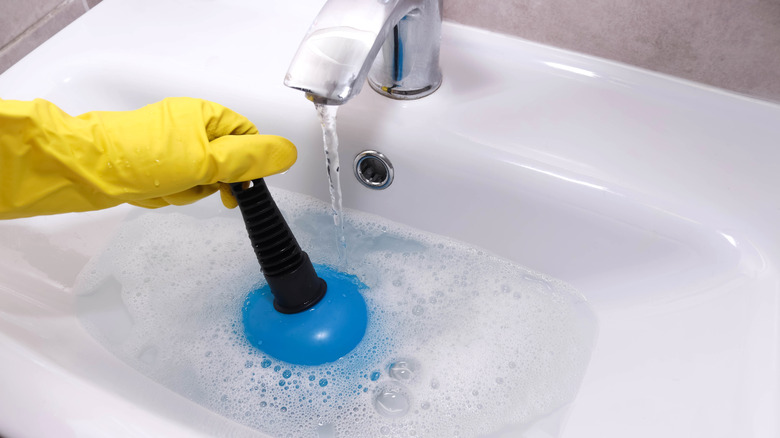 A gloved hand using blue plunger to unclog sink drain