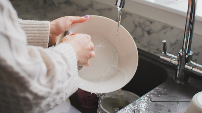 person in white knit sweater and pink nail polish washing white dish in the sink
