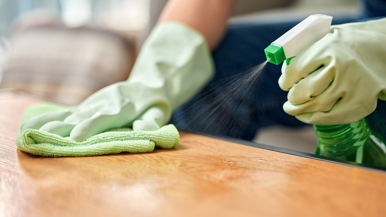 A person spraying a cleaning solution onto a wood table while wiping with a green microfiber cloth