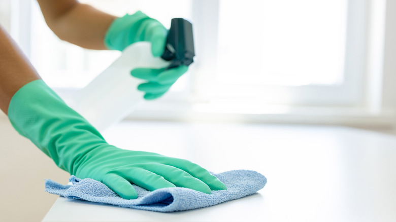 Someone in green gloves takes to cleaning the surface of a white table with a spray bottle and rag.