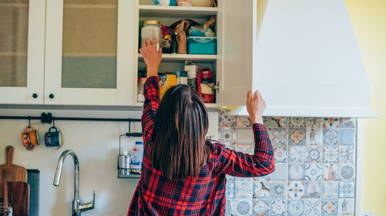 A woman in a red plaid shirt reaching for a jar in a kitchen cabinet