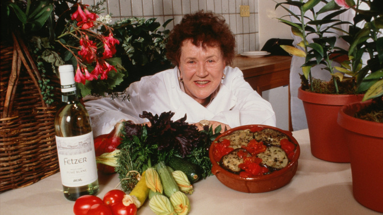 Julia Child facing the camera with wine, fresh vegetables, and a cooked dish