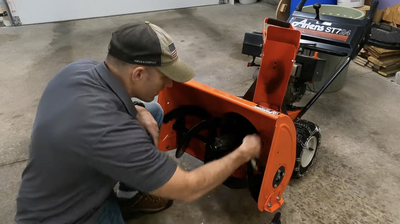 A man cleans debris from his snow blower before using it to clear standing water in his yard.
