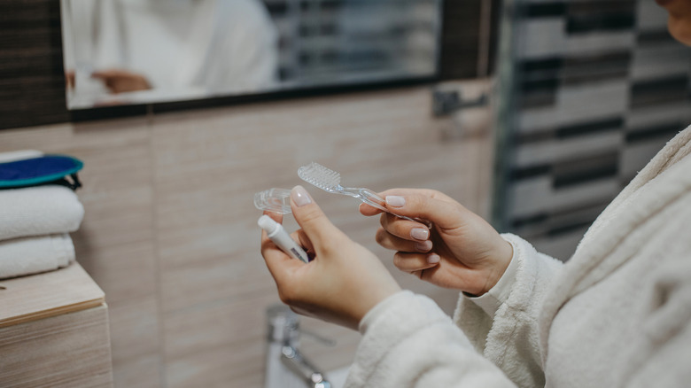 Person holding a toothbrush and toothpaste in front of bathroom mirror