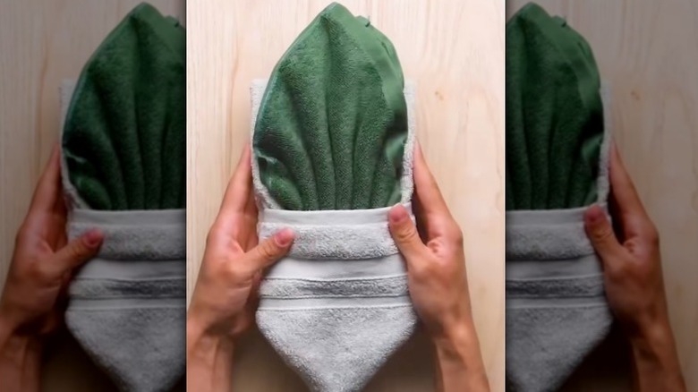 Hands hold the sides of a green hand towel tucked inside a white bath towel, both folded hotel-style.