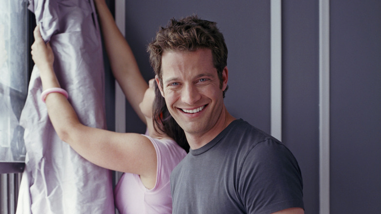 A headshot of Nate Berkus smiling at the camera with another person hanging curtains behind him