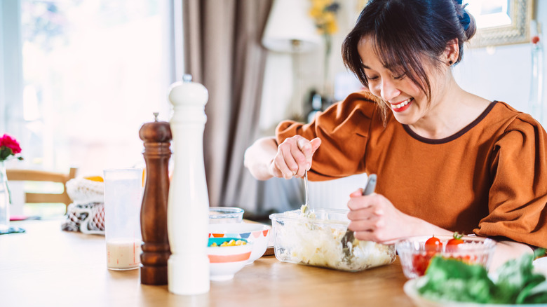 A woman smiles as she looks down at the food she is preparing as a mismatched salt and pepper shaker set sit on the table.