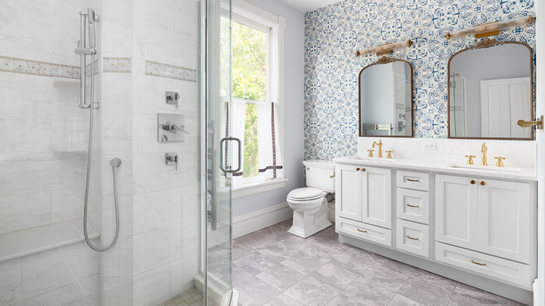 A modern bathroom with a walk-in shower, white cabinets, duel sinks and mirrors, and patterned blue and white wallpaper