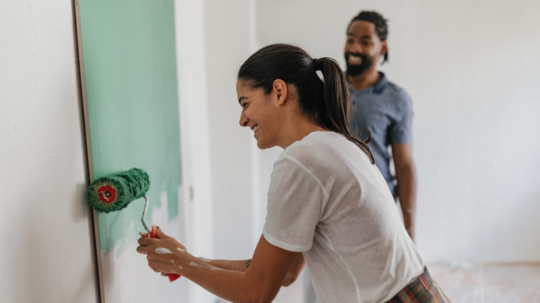 A woman with a paint roller covering a white room with a jade green color while a man stands behind watching