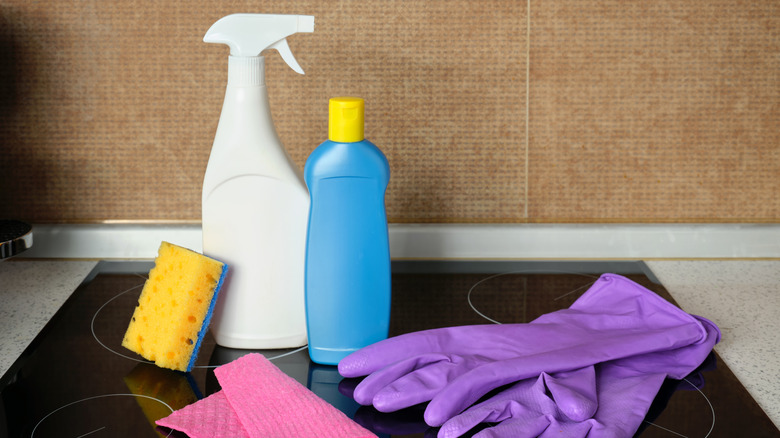 Household cleaning supplies on a kitchen stovetop, including spray bottles, a sponge, cleaning cloths, and purple rubber gloves.