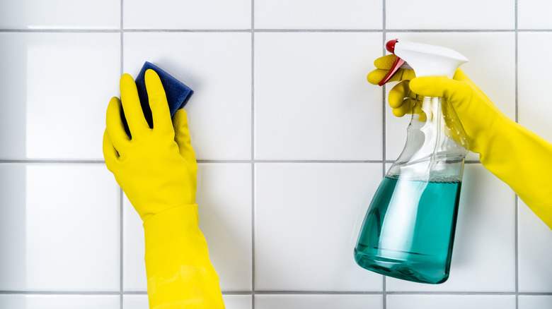 A person wearing yellow rubber gloves holds a bottle of teal cleaning solution and a sponge against a white tiled wall with light gray grout