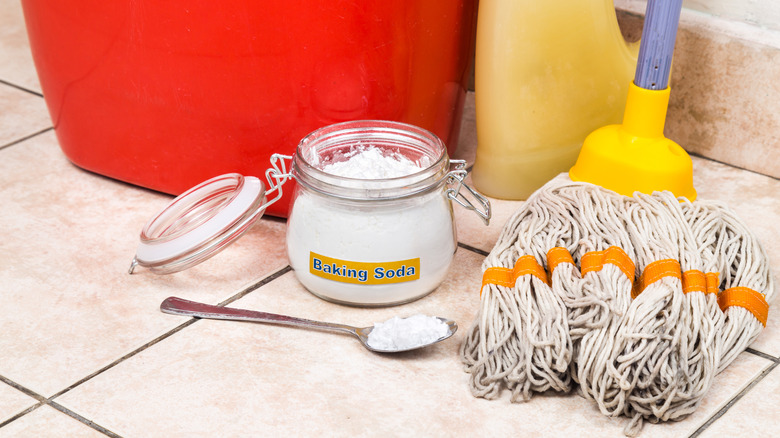 A jar of baking soda sits on a floor next to a bucket, a mop, and a bottle of cleanser.