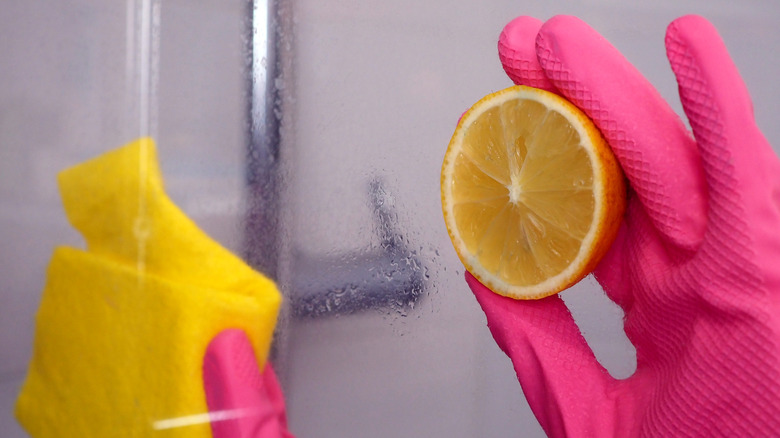 A person wearing rubber gloves rubs a lemon half and rag directly onto a shower door