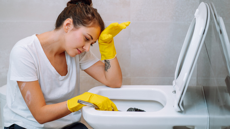 A woman in white shirt and yellow gloves struggles to clean the toilet bowl.