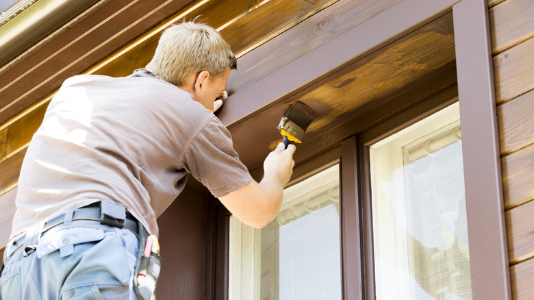 Worker applying brown paint with a brush to a wooden exterior wall above the window.