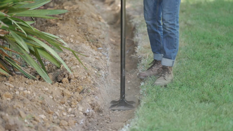 A person tamping down retaining wall base between a garden and a green lawn