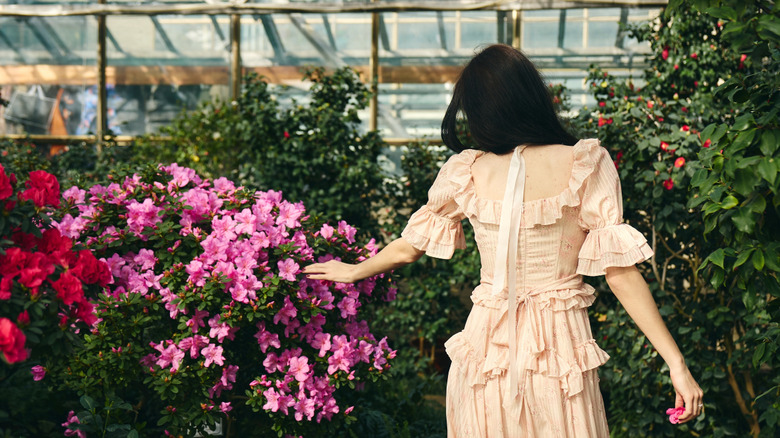 A woman in a romantic white dress walks through a greenhouse examining azaleas