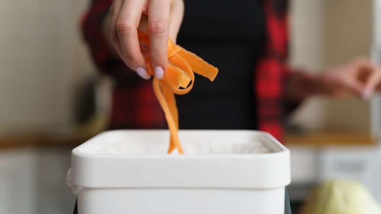 A woman's hand dropping scraps of carrot into an open white compost bin