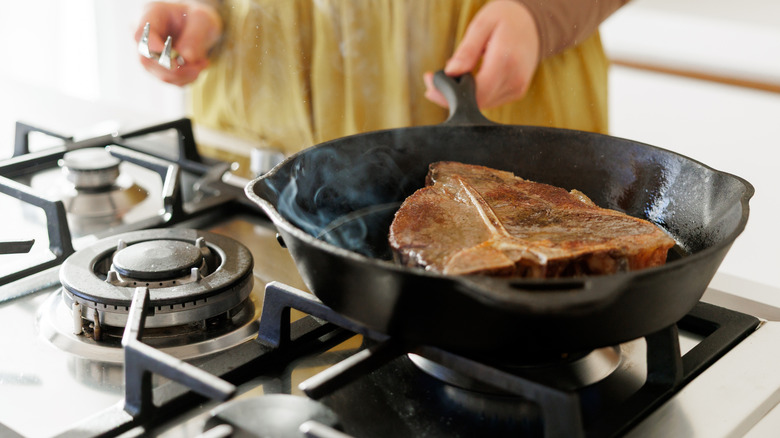 A person cooking a steak in a cast iron pot on a gas stove