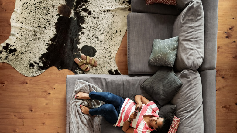 High angle view of woman lying comfortably on a grey sofa and using a mobile phone in her living room relaxing and enjoying some down time