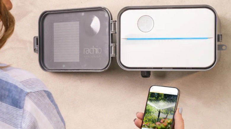 A hand holding a phone with the rachio app open in front of a sprinkler controller
