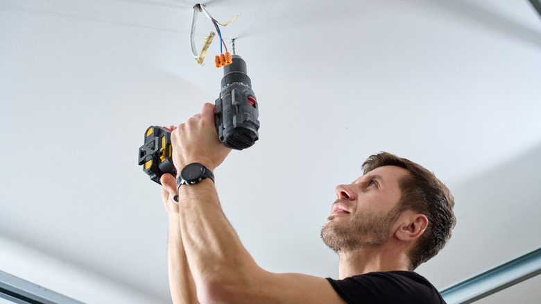 Man using a drill on a garage ceiling next to a hole with wires sticking out