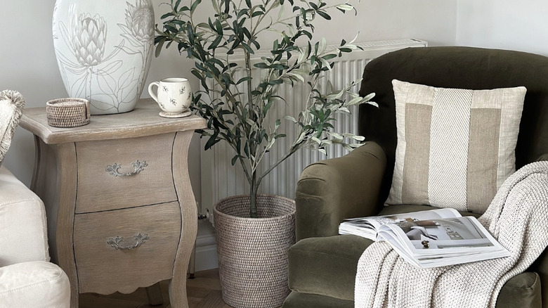 A living room corner decorated with modern cottage elements including a green armchair with throw blanket, a tall plant, and a wood side table