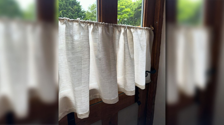 A beige cafe curtain hanging in a dark wood frame window with trees outside.