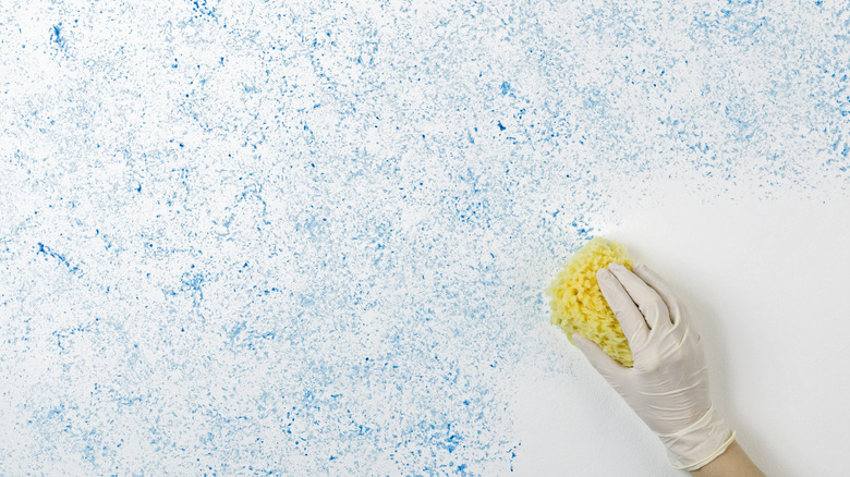 Sponge-paint texture being applied to a wall using a yellow sponge in someone's hand.