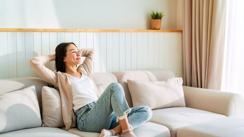 A woman sits on a couch in a warm living room with taupe curtains