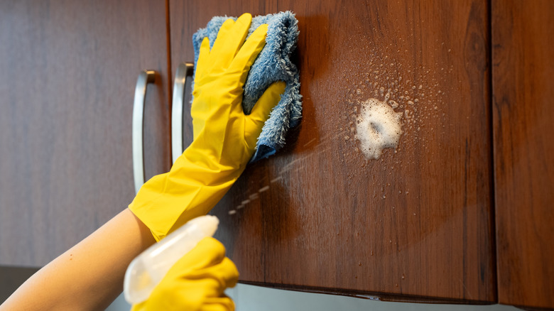 Hands in yellow rubber gloves spraying a wooden cabinet with one hand and holding a washcloth in the other